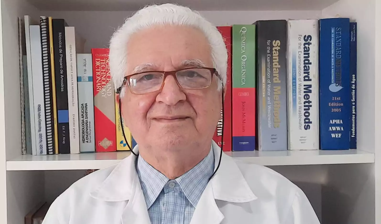 Doutor Samir Barcha, diretor técnico, com jaleco e óculos. Foto de perfil, em frente à sua prateleira de livros.